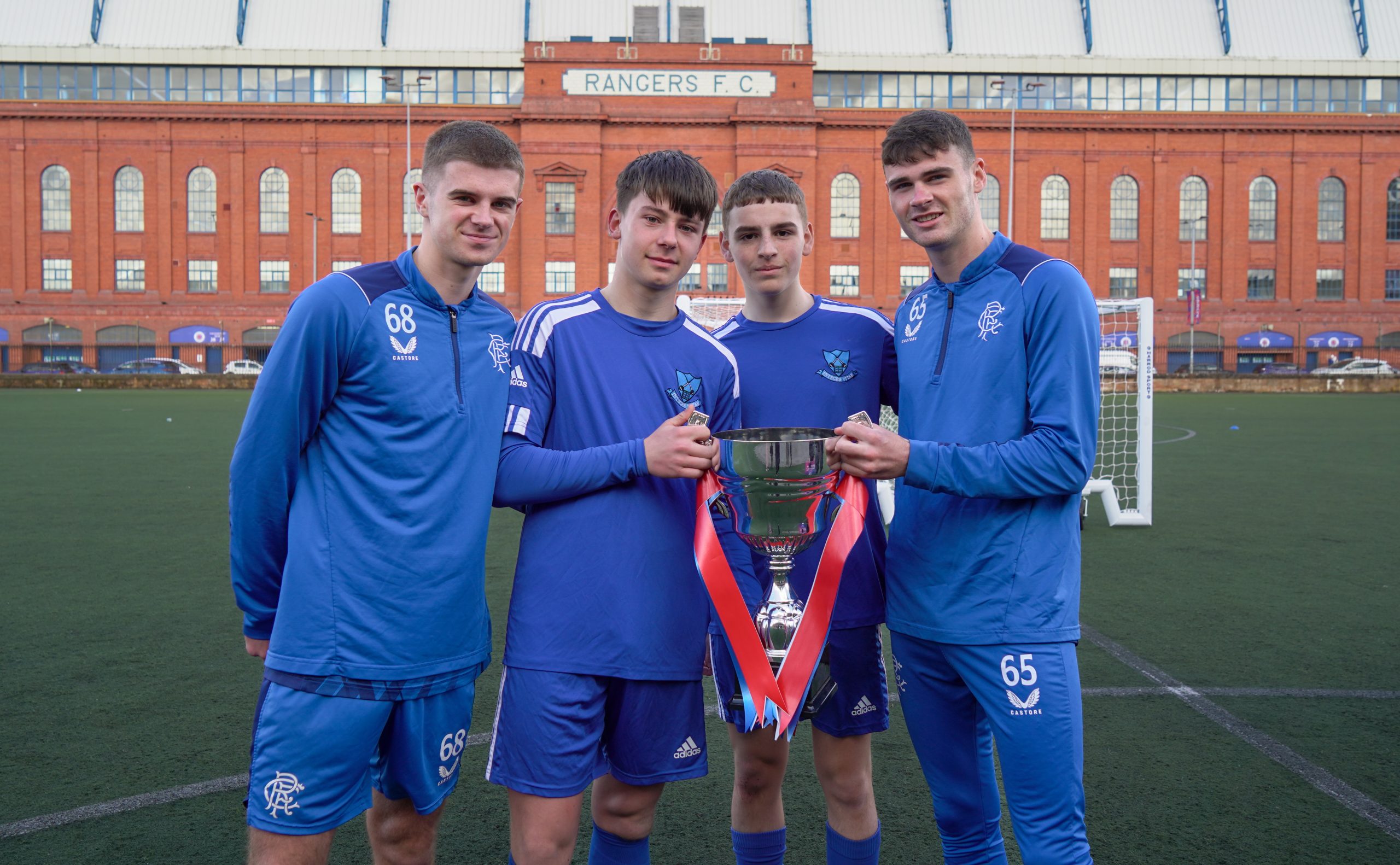 Two young male football players holding a silver trophy accompanied by two senior players either side