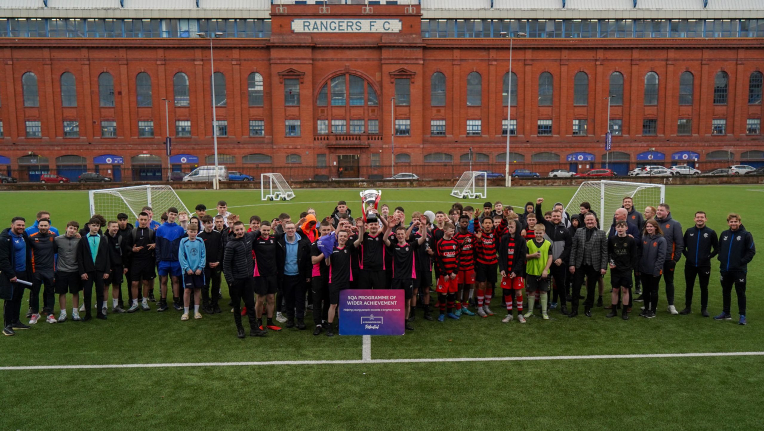 A big group of young people celebrating with a trophy in front of Ibrox