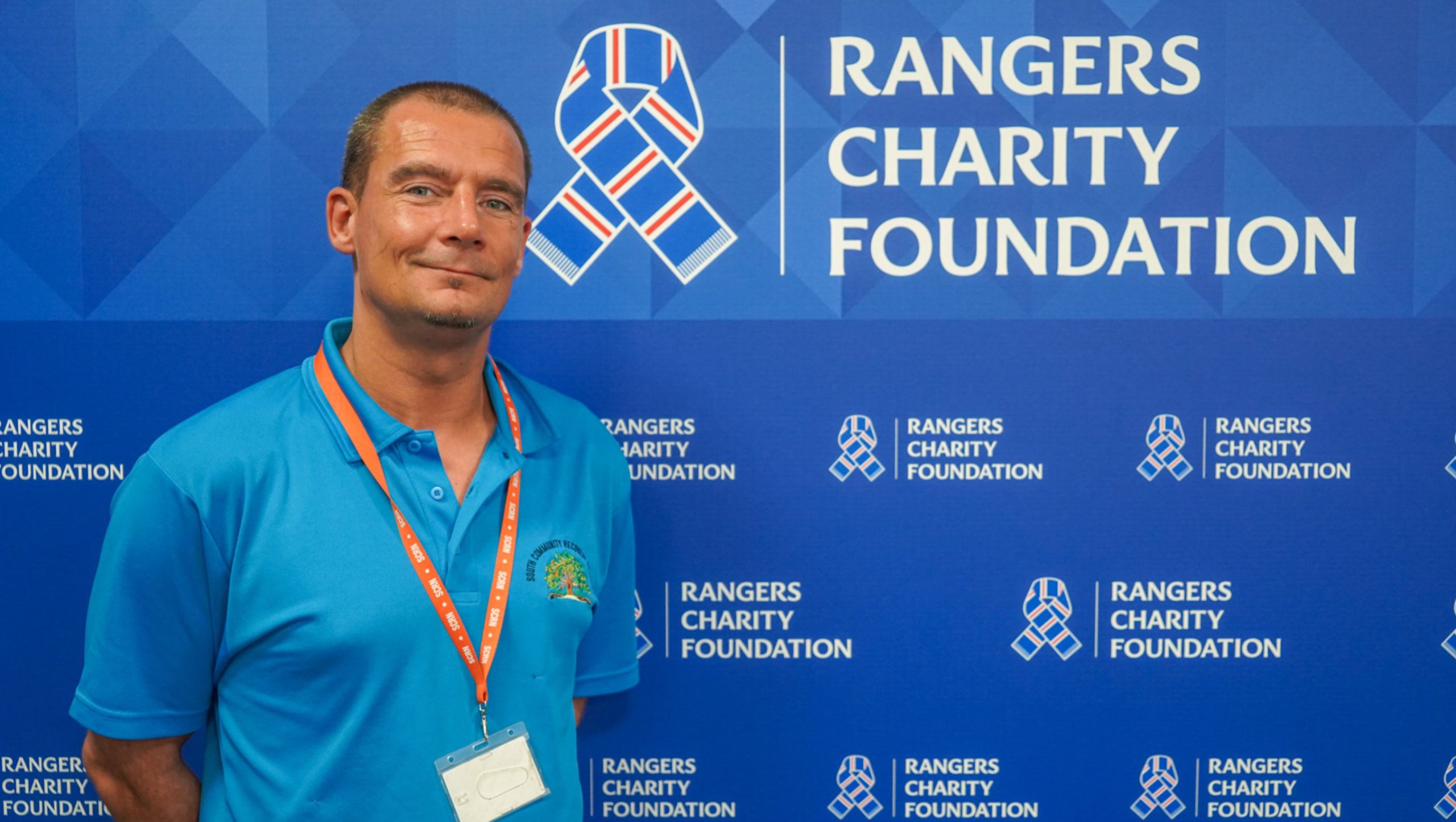 A smiling man standing in front of a Rangers Charity Foundation backdrop
