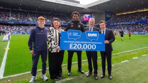 Rangers players Yilmaz, Fernandes and McCallion join Foundation CE Connal Cochrane and SAMH's Hazel McIlwraith on the pitch at Ibrox with £20K prop